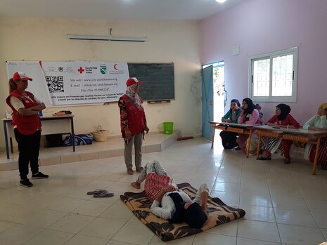 Eine Frau liegt auf einer Decke auf dem Boden. Daneben steht eine Lehrerin und es sitzen mehrere Schülerinnen im Hintergrund.